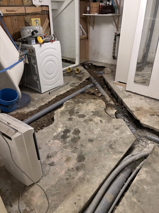 Renovering av källare med synliga pex-rör i betonggolv, vit tvättmaskin, verktyg och blå hink i bildens bakgrund.