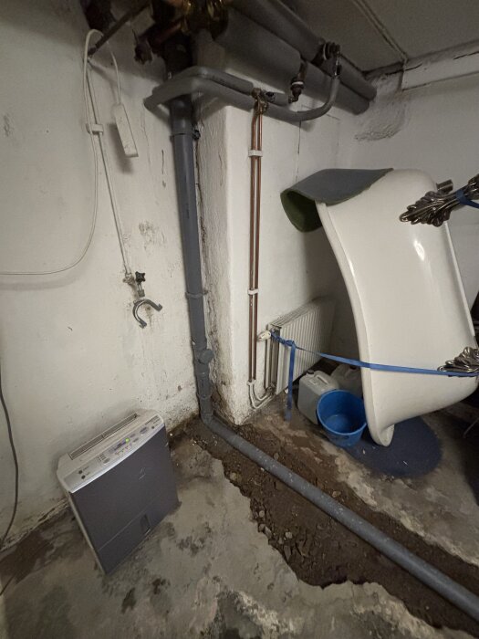 Källaren med synliga avloppsrör, en gammal vit badkarsdel, och en blå hink; nytt PEX-rör i betongplattan.