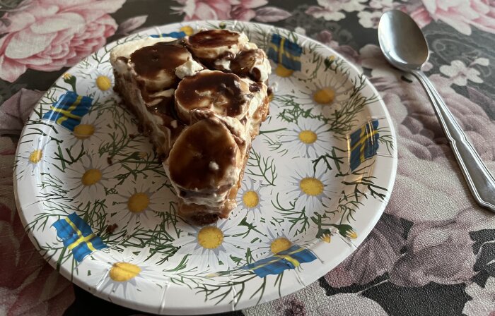 En bananoffeepajbit på papperstallrik med blom- och flaggmönster, bredvid en sked.