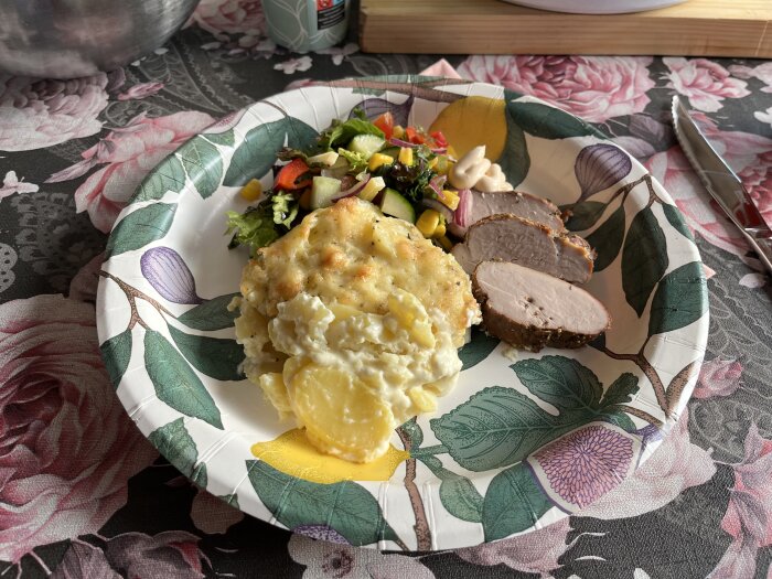 Pappersassiett med rökt fläskfilé, potatisgratäng och sallad på duk med blommönster.