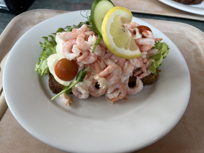 Räkmacka med bröd, sallad, räkor, citron, gurka, tomat och majonnäs på en tallrik.