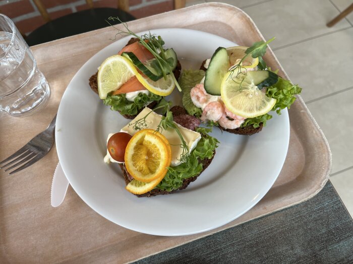 Tre smörrebröd på en tallrik med lax, citron, räkor och ost, garnerade med sallad, tomat och apelsin.