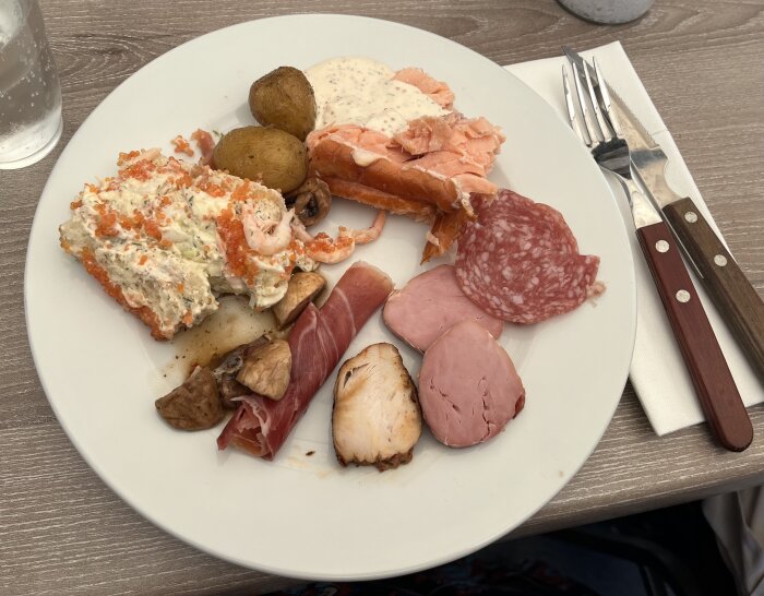 Tallrik med mat: räkmacka, lax, charkuterier, småpotatis och svamp. Förrätt från söndagsbuffé med olika köttsorter och röror.