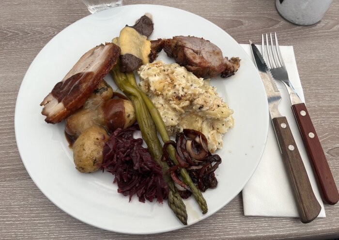 Tallrik med kött, potatisgratäng, grönsaker och sås från söndagsbuffé. Bestick och servett ligger bredvid på bordet.