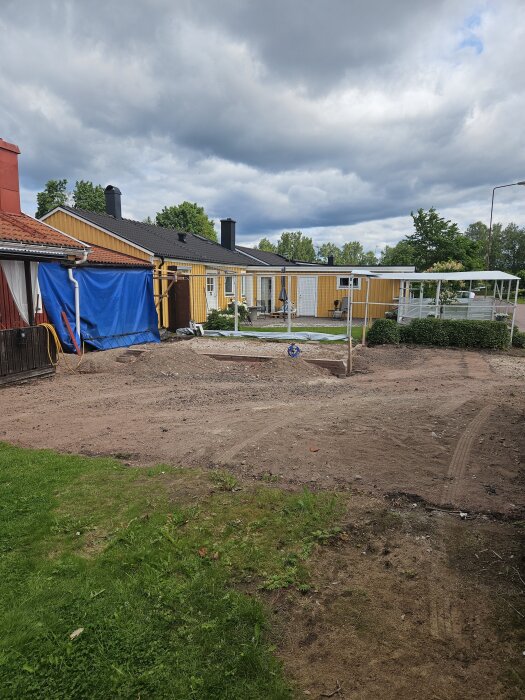 Tomt med grus och gräs framför ett gult hus, med byggmaterial och presenningar i förgrunden under molnig himmel.