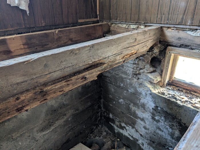 Gammal träbjälke med skador och mögel i ett rum, visar en skadad vägg och ett smutsigt hörn med litet fönster upptill.