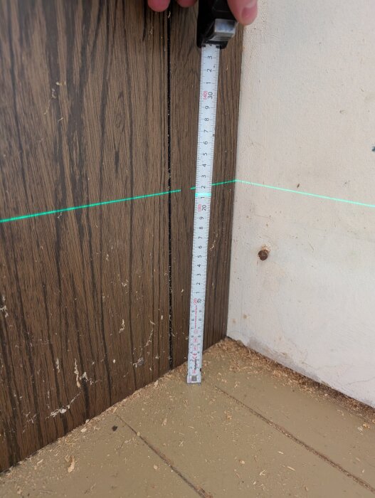 En laserlinje visar nivåskillnader på en vägg med träpanel och golv, med ett måttband som mäter avståndet från golvet till laserlinjen.