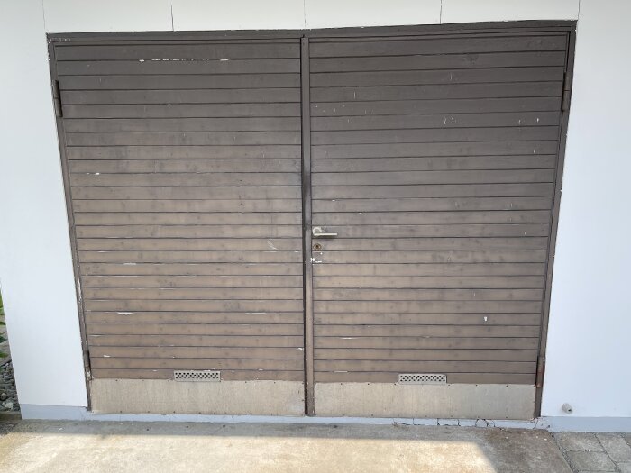 Två gamla garageportar med sliten brun färg, stående mot en vit vägg, med ett lås i mitten och ventilationsgaller längst ner.