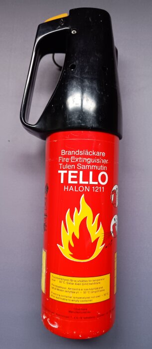 Röd halonsläckare med texten "TELLO HALON 1211" och en stor gul eldikon på framsidan.
