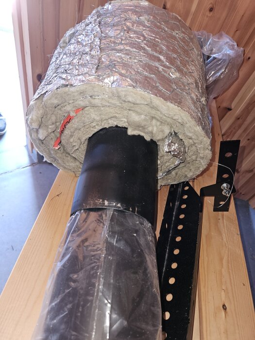 Oisolerat rökrör med isolering, omgiven av metall, placerad på träbord, redo för installation i trävägg.