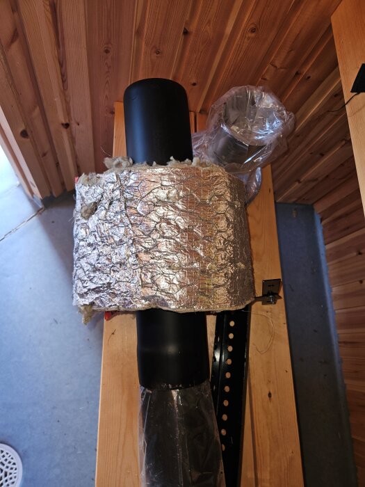 Oisolerat svart rökrör med silverfärgad isolering placeras genom en trävägg. Plast och metallkomponenter syns i bakgrunden.