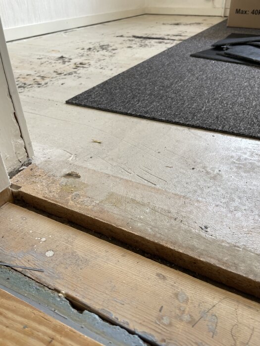 Närbild av ett rum med en delvis borttagen matta, blottad underliggande träskiva och fläckar av limrester.