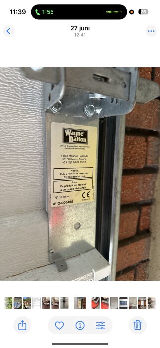 Etikett från garageportsmärket Wayne Dalton på en metallbeslag mot en tegelvägg. Etiketten visar företagsinformation och serienummer.