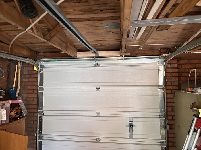 Insidan av en garageport med träbjälkar i taket och tegelväggar. Porten har ett metallstyrsystem och är stängd. Verktyg syns på sidan.