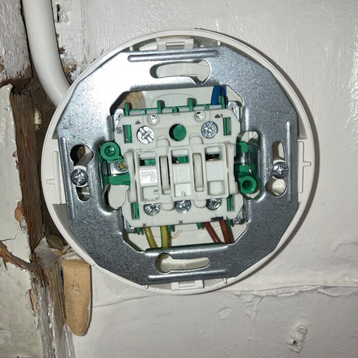 Elektrisk kopplingsdosa med grön, blå och brun ledning synliga, ansluten för taklampa. Bakgrund av vitmålad vägg med trädetaljer.