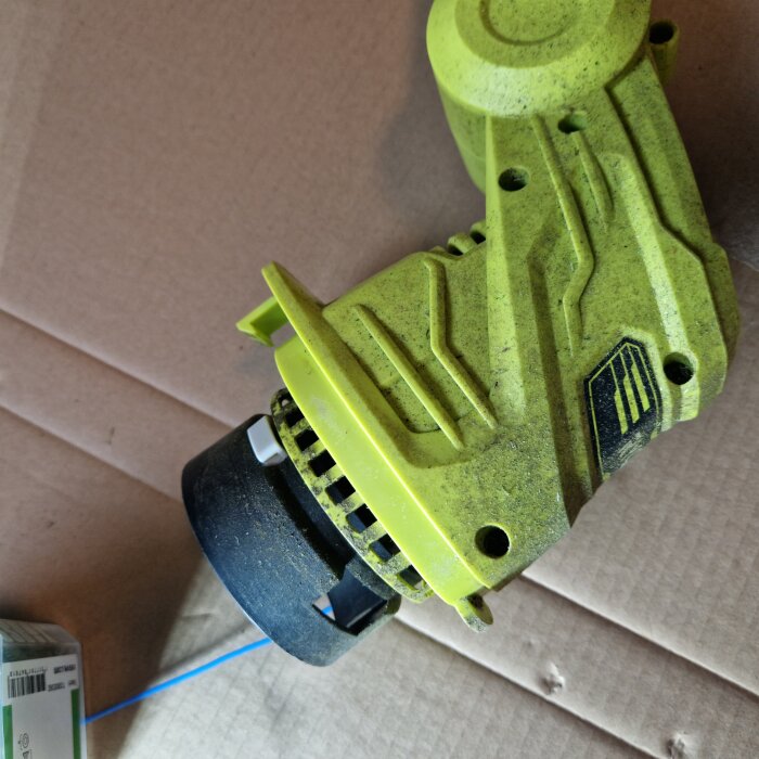 Gul Ryobi OLT1832 trimmer med fastnad tråd vid spolen, liggande på en kartongyta.