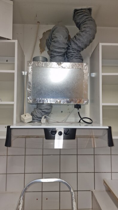 Spiskåpa i kök under installation, med synliga ventilationsrör och kablar. Ovanför finns ett metalliskt aggregat med anslutande slangar.