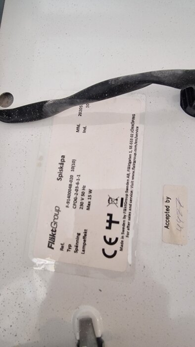 Etikett på en spiskåpa från FläktGroup med specifikationer som typ, spänning och lampeffekt, delvis täckt av ett svart band.