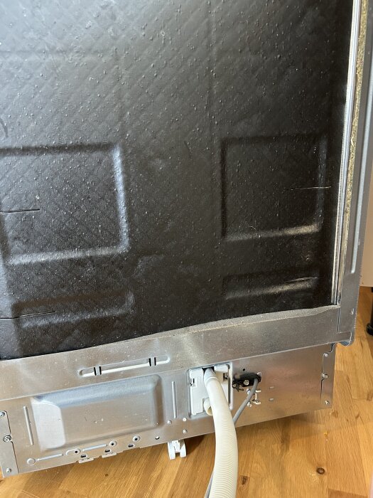 Baksidan av en diskmaskin med avloppsslang ansluten, viss metall, trä underlag och några skruvar synliga.