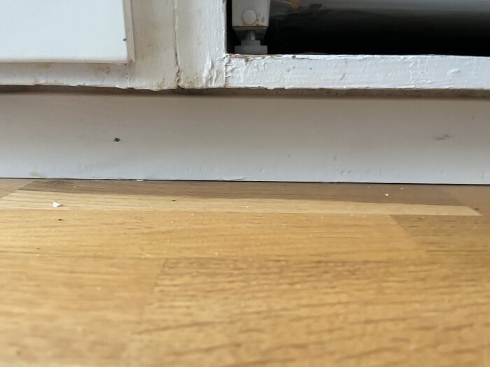 Närbild på kökssockel och golv, med fokus på ett slitet skåp på sockeln, för att visa potentiell förändring inför kylskåpsbyte.