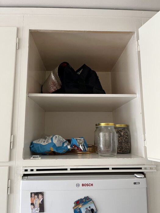 Överskåp med två hyllor, fyllda med bland annat en kylväska, chips, glasburkar och stående ovanpå ett kylskåp av märket Bosch med magneter.