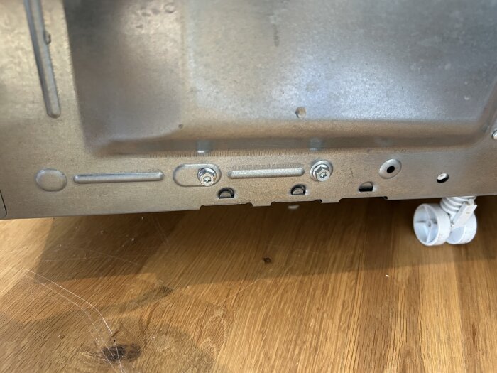 Baksidan av en maskinkaross som visar metallskruvar och kabeldragning på ett trägolv.