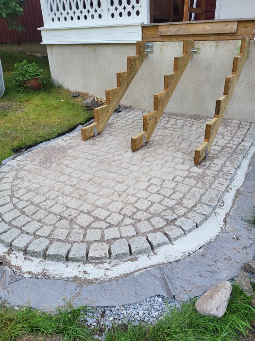 Stegkonstruktion över ny stenläggning framför veranda med luckra jordytor och intention att så gräs vid stenarnas kant.