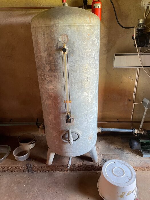 Gammal hydrofor med mätare och ventil, placerad i ett rum med tillhörande rörsystem och avlagda hinkar på golvet.