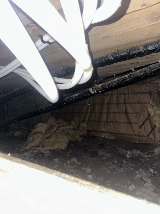 Vita vattenrör hänger i källartak med svart rörstöd. Isolering ligger löst på marken i en byggkällare.