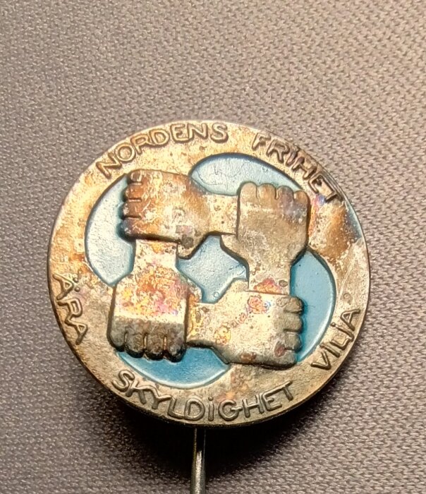 Nålmärke för Samfundet Nordens Frihet med fyra händer i ring och texten "Nordens Frihet Ära Skyldighet Vilja" i en cirkel.