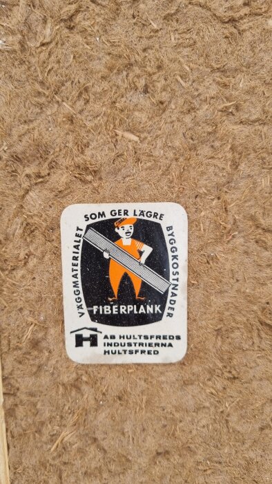 Etikett på fiberplank med texten "Som ger lägre byggkostnader", med en illustration av en byggarbetare som håller en planka.