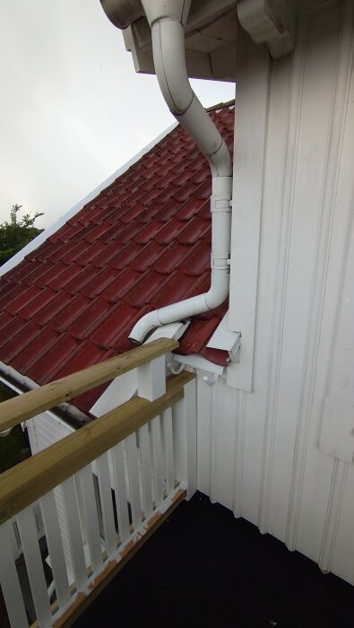 Vit stupränna som leder vatten ner från ett rött tegeltak, ansluten till en ny träbalkong.