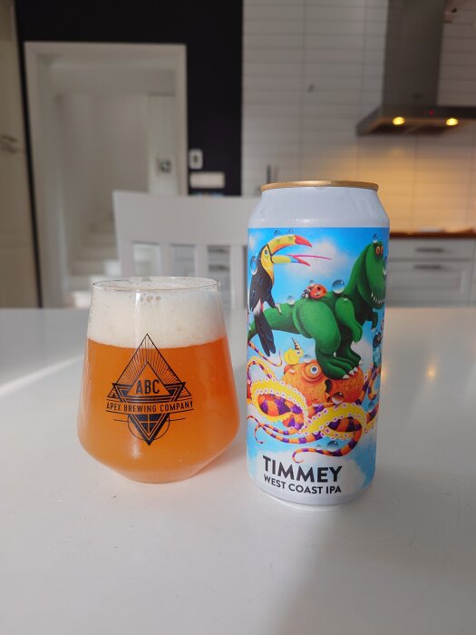 En burk Timmy West Coast IPA med färgglad etikett bredvid ett glas med öl på ett vitt köksbord.