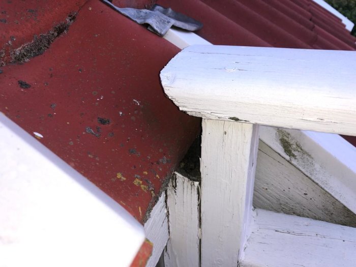 Vitmålat räcke möter röd takpanna på ett hus, med en tydlig fog ovanpå balkongen. Stuprör och hängränna saknas.