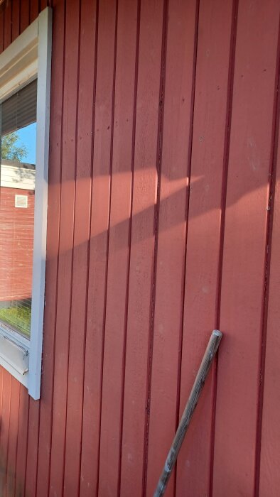 Röd fasadpanel med trästruktur och fönster, delvis skuggad, med trästolpe lutad mot väggen.