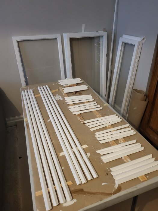 Vita fönsterramar och lister ligger på ett bord, redo för montering.