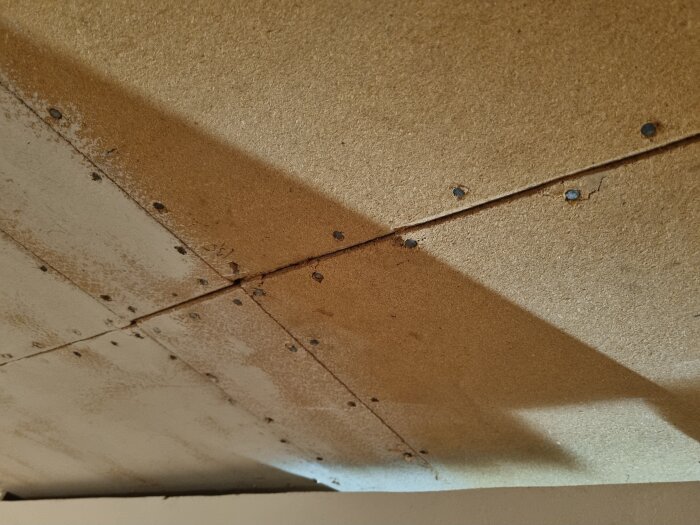 Bilden visar spånskivor som tak i en källare, med glassfiberull isolering ovanpå. Spånskivorna är skruvade ihop och visar viss fukt eller missfärgning.