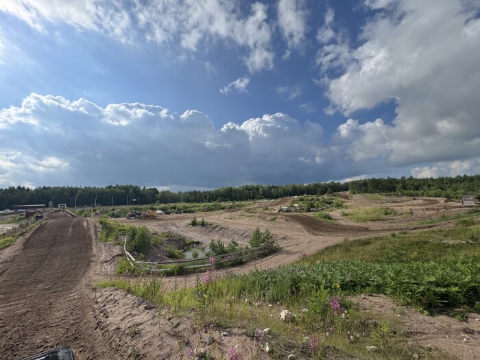 Motocrossbana under solig sommardag med blå himmel och vita moln, omgiven av skog och grönska.