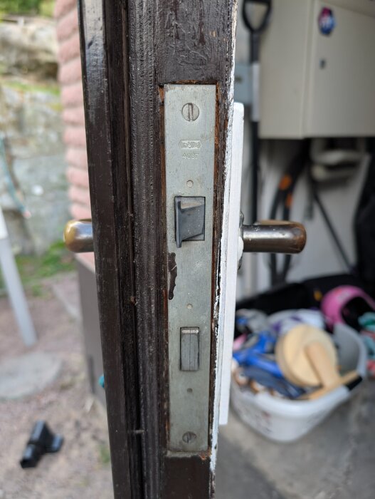 Närbild av ett dörrlås i en träkarm med synlig skruv och handtag. Detaljerad fokus på låsets konstruktion och omgivningen i bakgrunden.