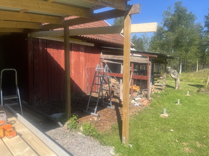 Byggarbete på baksidan av ett rött uthus med trästolpar, stegar och byggmaterial, samt en gräsplan och träd i bakgrunden.