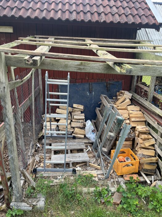 Uthus med takstomme av trä, lådor med ved, stege och en orange plastback på marken framför en röd träpanel och ståltrådsstängsel.