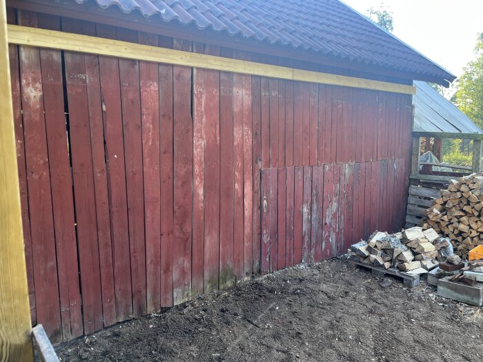Röd trävägg med tak, ved staplad intill, del av ett renoveringsprojekt på baksidan av ett uthus.