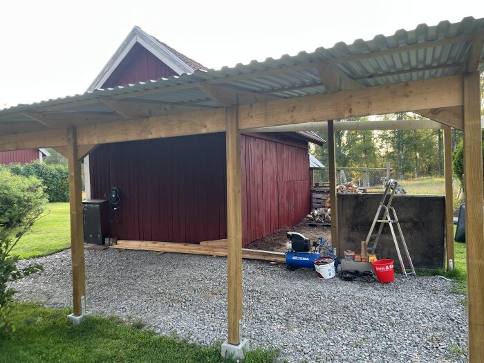 Carport med trästruktur och tak i färdigställande. Rödmålat uthus i bakgrunden, stege och byggmaterial på marken. Grönskande omgivning.