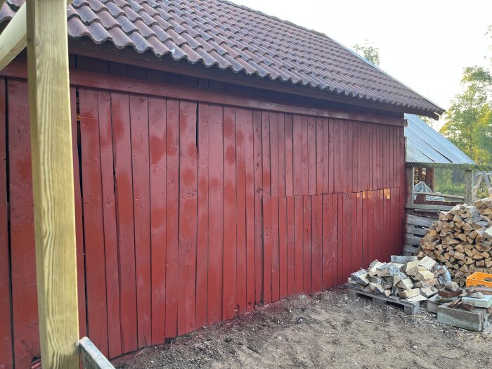 Röd uthusvägg med ny målning, staplad ved vid sidan och en ny trästolpe i förgrunden, del av ett pågående renoveringsprojekt.