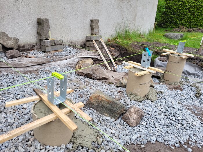 Betongplintar med byggbeslag monterade i en grusad yta, förberedelser för ny trappa utanför ett hus.