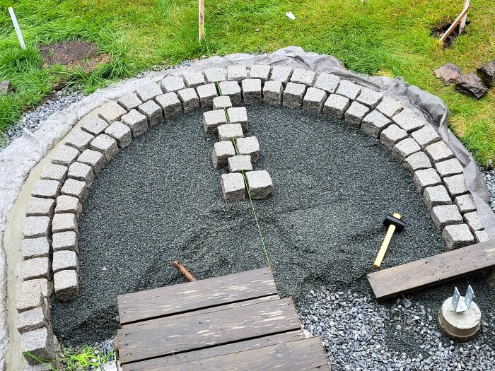 Byggarbetsplats med halvfärdig gatstenlagd trappa, synlig gummiklubba och plankor, omgiven av grus och gräs.