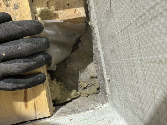 Hand med handske lyfter träplanka, exponerar isolering och plast över betong och stenkross i källargolv.