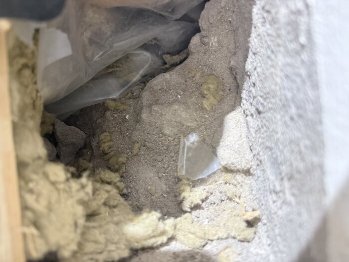 Isolering och stenkross synliga under ett uppsågat trägolv i ett källarrum, med plast och betongfragment i bakgrunden.