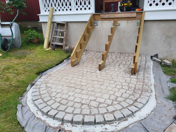 Trappa under uppbyggnad med trästeg och nylagd gatsten på marken, bredvid ett vitt räcke och grönytor.