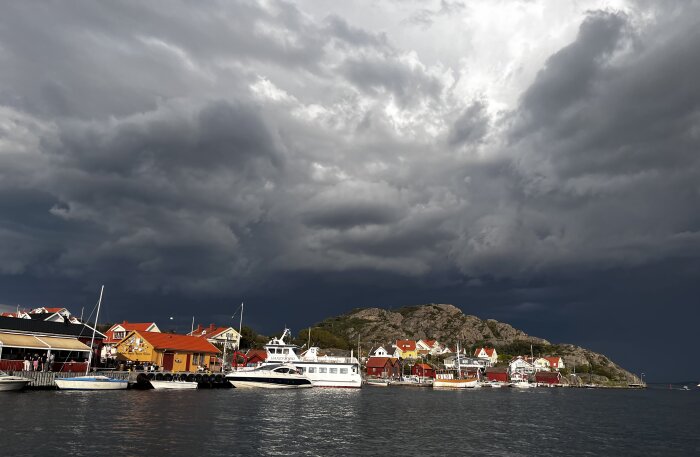 Mörka moln över en kuststad med båtar och färgglada hus.